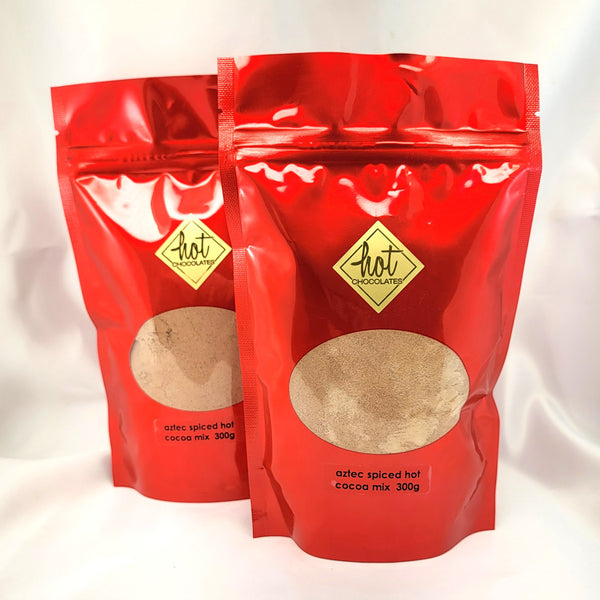 Two glossy red stand-up pouches of Hot Chocolate Aztec Spiced Artisan Hot Cocoa Mix 300g, displayed against a white satin fabric background.