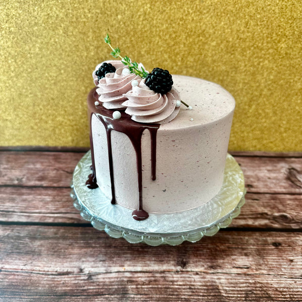 A pretty layer cake with light purple icing decorated with chocolate and blackberries and filled with blackberry compote and mascarpone custard, displayed on a cake stand.