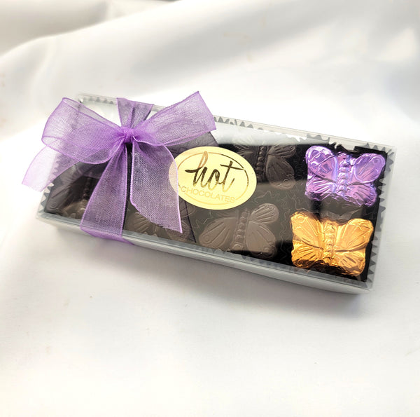 A rectangular gift box of single origin Colombian chocolates tied with a purple organza ribbon and a round gold label reading 'Hot Chocolates'. The box is clear plastic, showing several dark chocolates inside, along with two butterfly-shaped chocolates in purple and gold foil wrappers, set against a white fabric background.