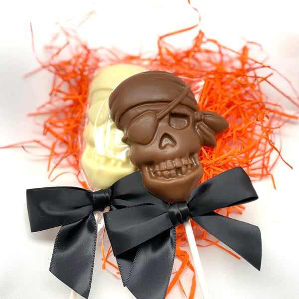 2 Chocolate Pirate Lollipop Suckers designed to look like a skull, with a black bow and orange shred around it, one in milk chocolate and one in white.