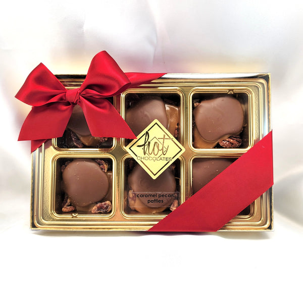6 boxed caramel pecan patties in a gold gift box tray, tied with a large red satin bow and ribbon, on a white background. A central label reads 'hot CHOCOLATES'.
