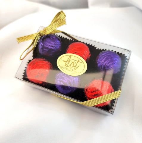 A clear rectangular gift box containing six wrapped artisan chocolate covered cherries in red and purple foil, tied with a gold ribbon and a gold oval label reading 'Hot Chocolates', photographed on a white fabric background.