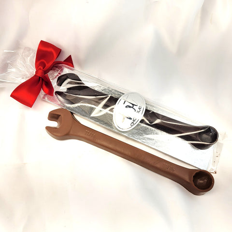 A chocolate wrench gift featuring a large milk chocolate wrench in the foreground with 11/16 visible on the neck and a packaged dark chocolate wrench tied with a red ribbon in the background, on a white fabric backdrop.