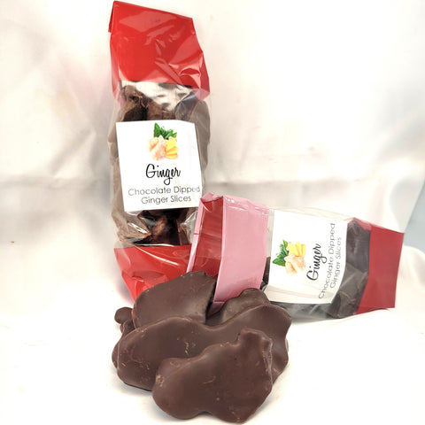 Two packages of chocolate dipped ginger slices with loose pieces in front, on a white fabric background. The packages are red and clear, showing dark and milk chocolate options, with labels reading Ginger Chocolate Dipped Ginger Slices.