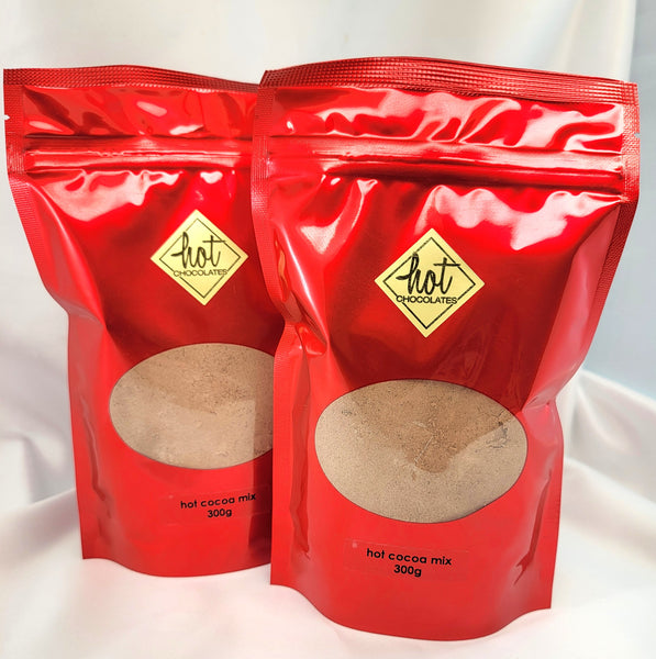 Two red stand-up pouches of Hot Chocolate artisan traditional hot cocoa mix, 300g each, with a clear oval window showing the cocoa powder inside, set against a white draped fabric background.