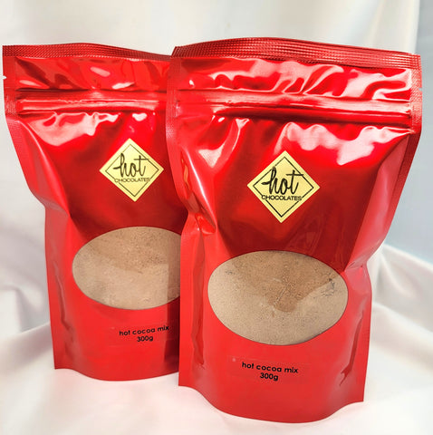 Two red stand-up pouches of Hot Chocolate artisan traditional hot cocoa mix, 300g each, with a clear oval window showing the cocoa powder inside, set against a white draped fabric background.