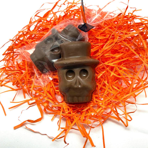 Two skull-shaped chocolate Mexican Hat Calavera candies wrapped in clear cellophane, displayed on bright orange shredded paper against a white background.