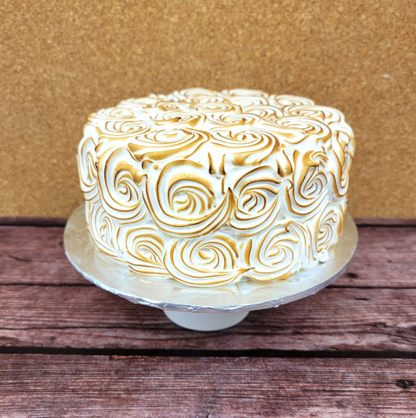 A round no gluten added lemon cake with white and golden-brown toasted meringue swirls on top and sides, displayed on a silver cake stand against a brown corkboard and wooden surface.