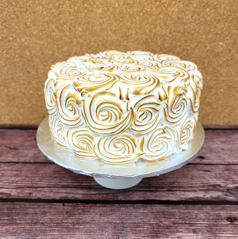 A round no gluten added lemon cake with white and golden-brown toasted meringue swirls on top and sides, displayed on a silver cake stand against a brown corkboard and wooden surface.