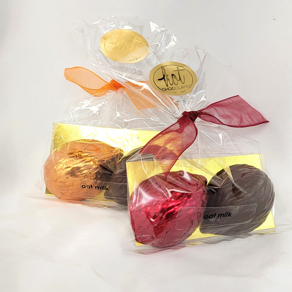 Two packaged chocolate hedgehogs on a white background. The front package contains dark chocolate, dairy free oat milk hogs with one wrapped in red foil, tied with a burgundy ribbon.