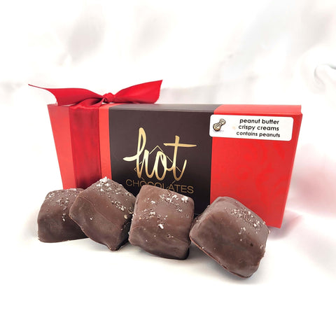 A red and brown gift box of hot chocolates tied with a red ribbon, with four chocolate pieces placed in front on a white background. A small label on the box reads 'peanut butter crispy creams contains peanuts.