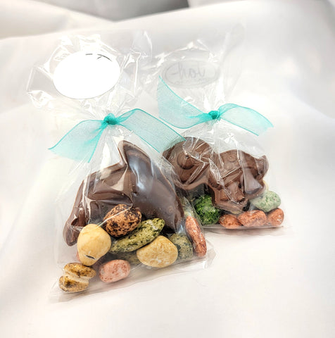 Two clear cellophane ocean-themed treat bags tied with aqua ribbon, filled with assorted chocolates and colorful candy rocks photographed on a white surface.