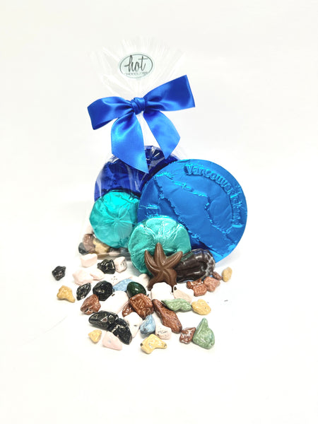 A colourful collection of goodies in the Vancouver Island chocolate Beachcomber bag, including: a large Vancouver Island chocolate coin and a chocolate sand dollar, both foiled in ocean colours. Plus chocolates sea creatures and edible rocks on a bed of white.