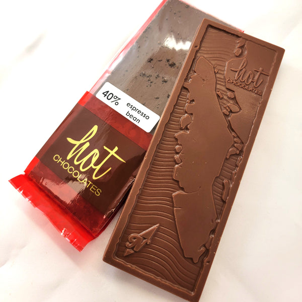 An artisan chocolate bar packaged and an unwrapped chocolate bar on a white background. The package is dark red with gold script reading 'hot CHOCOLATES' and a white label that says '40% espresso bean'.