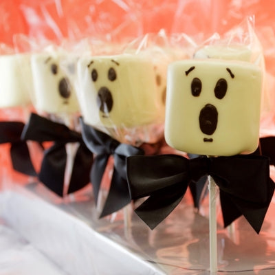 A collection of Chocolate Dipped Marshmallow Ghost Pops with scared faces, in clear bags with a black bow on a peach coloured background.