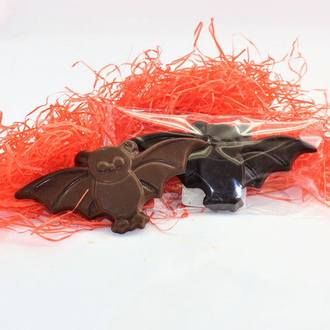 2 Solid Chocolate Halloween Bat Treats with a glossy finish, placed on a surface with orange shred. The candy is wrapped in a clear plastic packaging.