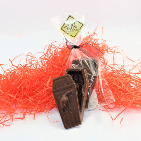 A Solid Colombian Milk Chocolate Coffin, shown with a removable lid to reveal a skeleton inside, bagged in a clear packaging, on a bed of orange shred. 