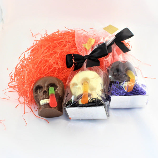 Three packages of Solid Colombian Chocolate Skulls & Candy shown in Milk, dark and white chocolate with gummy worms, each wrapped in individual clear bags, set against a white background with orange shred.