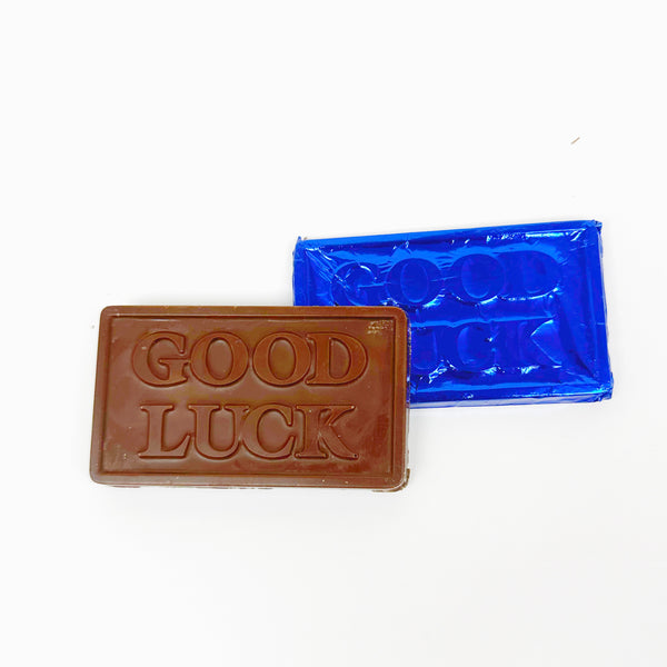 Two solid oat milk chocolate good luck bars with blue foil packaging, one bar is chocolate brown in color and the other is blue, both with the text 'Good Luck' printed on them.