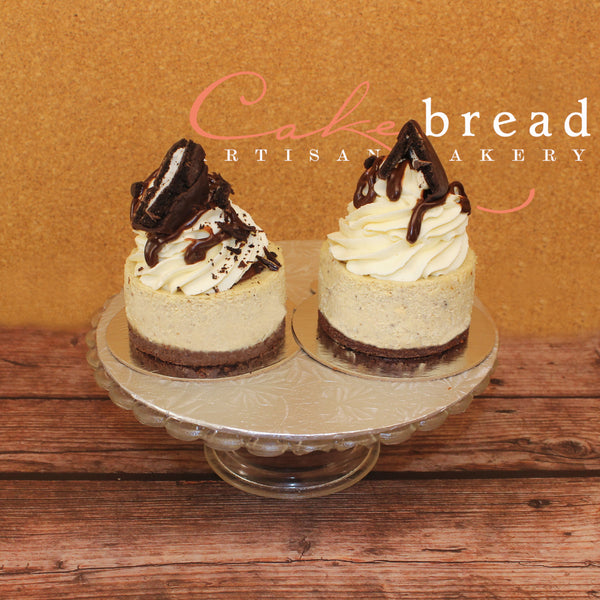Two mini New York-style cheesecakes with chocolate toppings and white frosting, presented on a plate with a swirl of chocolate on top.