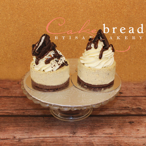 Two mini New York-style cheesecakes with chocolate toppings and white frosting, presented on a plate with a swirl of chocolate on top.