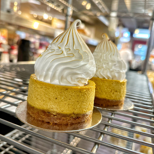 Two Mini New York-Style Pumpkin Spice Cheesecakes, with brown crusts, orange coloured cake and topped with toasted meringue on silver discs on a metal rack.