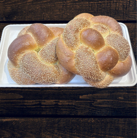 2 beautiful loaves of braided round Challah bread, finished with sesame seeds on a white tray and a wood background.