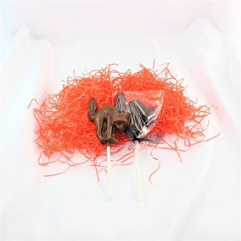 A Spooky Chocolate Cat Lollipop Sucker shown with an arched back and hissing mouth, placed on a white surface surrounded by orange shred.