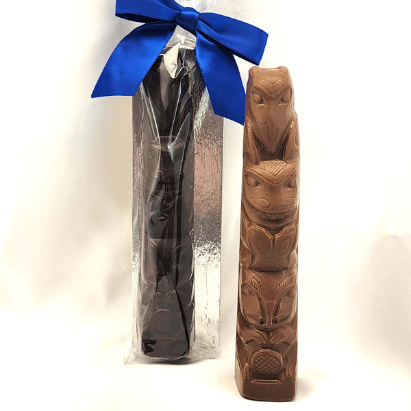 Two carved chocolate totem-style poles displayed side by side on a white studio background. The left object is wrapped in clear plastic with a bright blue ribbon bow at the top, and the right object is shown standing upright outside the packaging.