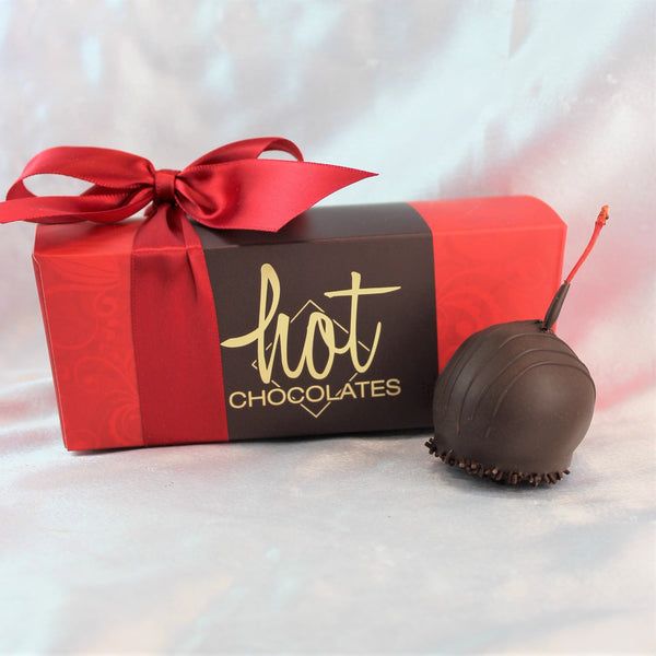 A bright red box in the back ground, with a red bow and a Brandied Cherry Dark Chocolate Truffle in front, with a stem poking out and sprinkles on the bottom.