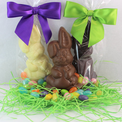 A solid chocolate Easter Bunny with jelly bellies, positioned in the center of a bed of green, with purple and green bows on the bags.