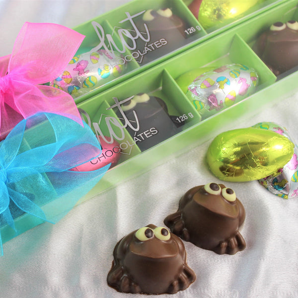 A box of Hot Chocolates branded chocolate frogs & Easter eggs, with a pink and blue decorative ribbon on a white surface.
