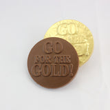 A chocolate coin with the phrase 'GO FOR THE GOLD!' embossed on it, placed next to a gold-foiled coin with the same embossing.