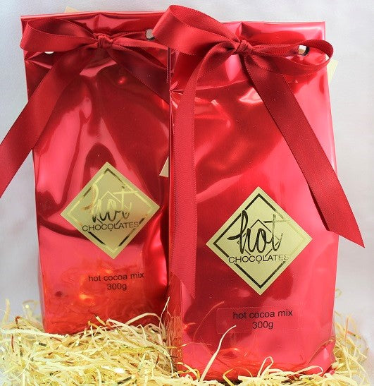 Two red packages of traditional artisan hot cocoa mix with a decorative bow on each, placed on a bed of straw. The brand 'Hot Chocolate' is visible on the packaging.