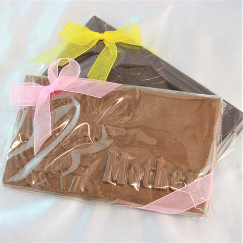 A large solid Mother's Day Chocolate Bar with 'Mother' written on it, wrapped in clear plastic with a pink ribbon and a dark chocolate version behind it with a yellow bow.