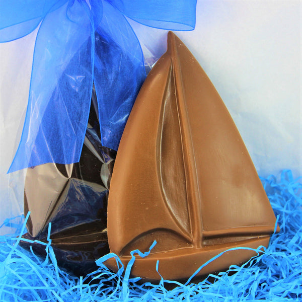 A solid chocolate sail boat made from milk chocolate in the front, with a dark chocolate one packaged and bowed in blue, on blue shred.