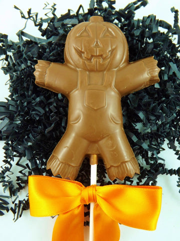 A Milk Chocolate Scarecrow Lollipop Sucker with a jack o lantern head and no hands or feet, on a white stick with an orange bow, on a bed of black shred.