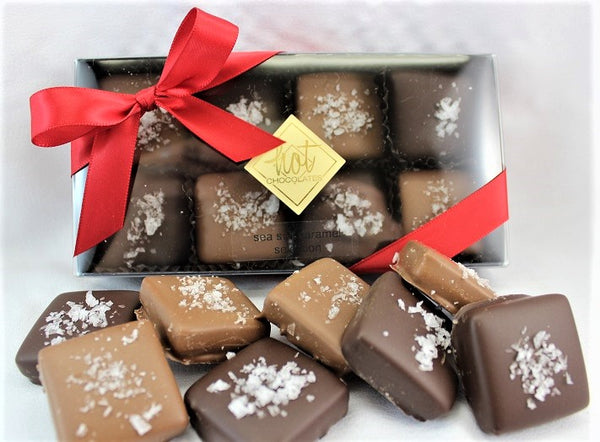 A beautiful see-through box with a red ribbon and a Hot Chocolates logo sticker, with artisan handcrafted sea salt caramel chocolates in the box and in front of it, on a white background.