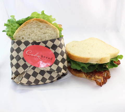 A gourmet BLT sandwich on fresh baked bread, wrapped in a checker paper with a Cakebread Artisan Bakery sticker, beside and unwrapped sandwich