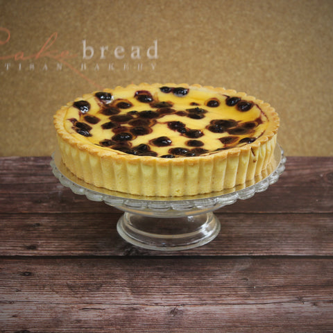 A baked yogurt tart with blueberries, with a beautiful golden tart shell on a glass cake stand with a rustic wood counter.