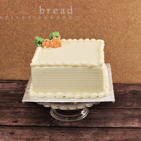 A rectangular classic carrot cake with cream cheese icing, decorated with a small carrot and leaf-shaped icing on top, placed on a glass cake stand.