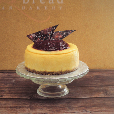 An artisan New York-style cheesecake with a seasonal fruit compote on top and fancy chocolate shards, presented on a glass cake stand.