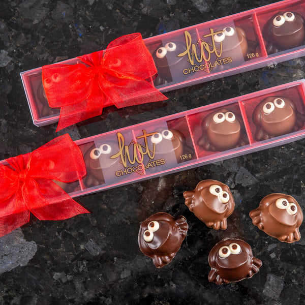A clear plastic box with six solid chocolate frogs, each with a smiling face, made from single origin chocolate. The box is adorned with a red bow at the top and a Hot Chocolates logo.