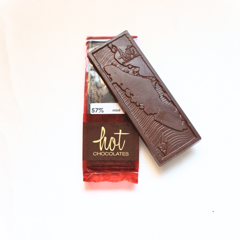 A dark artisan chocolate mint bar with a decorative pattern on its surface, placed next to its red packaging with the brand 'Hot Chocolates' visible.