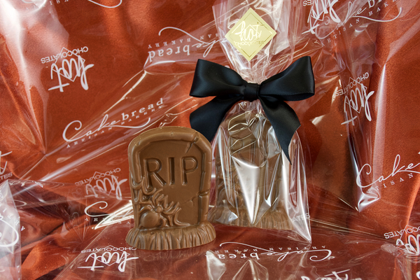 A solid milk chocolate tombstone shaped with RIP and a spider on the front, packaged in plastic and finished with a black bow, on a red background.
