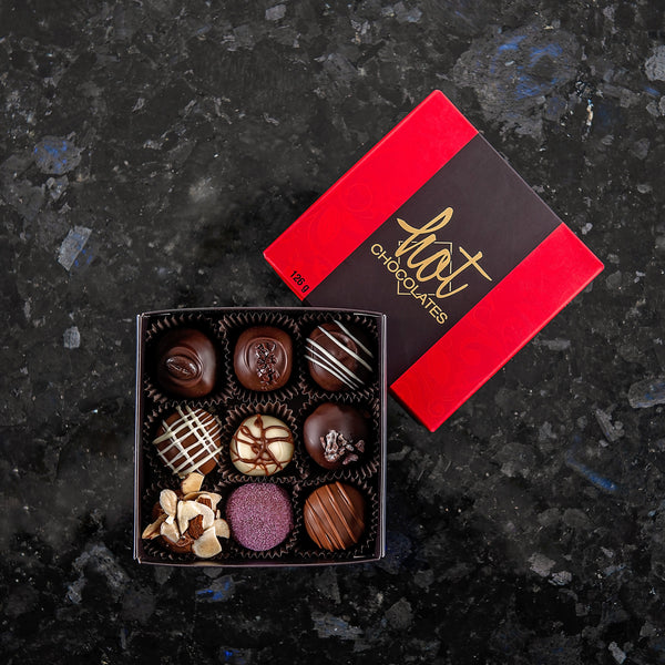 A box of 9 assorted hand-dipped chocolate truffles in a dark brown box with a red lid, displayed on a dark surface.