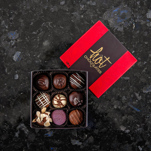A box of 9 assorted hand-dipped chocolate truffles in a dark brown box with a red lid, displayed on a dark surface.