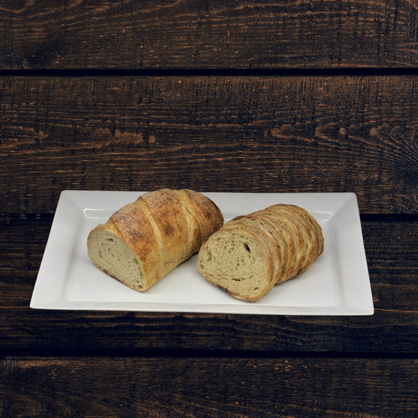 Two loaves of whole wheat rye sourdough bread, one sliced, on a white plate, placed on a wooden surface.