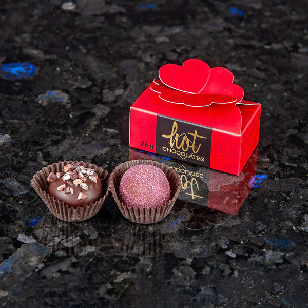 2 hand-dipped chocolate truffles sitting on a dark surface beside a red and brown chocolate box with a heart closure top. 