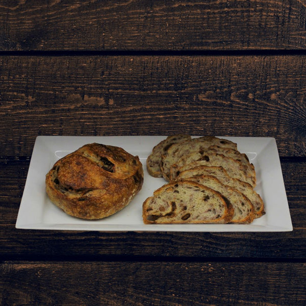 A plate with sliced and whole round loaves of fig & roasted walnut bread boule- one is a round with visible nuts and figs, and the other is a sliced loaf with similar inclusions, against a wooden background.